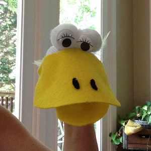 May include: A yellow felt puppet with white felt eyes and a black felt beak. The puppet is being held by a person's hand.
