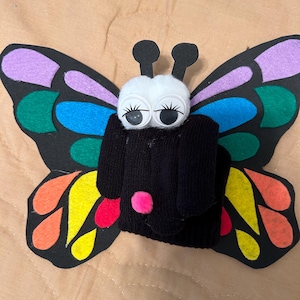 May include: A handmade butterfly craft made from a black sock, colorful felt wings, googly eyes, and a white pom-pom head. The wings feature vibrant colors like purple, green, blue, yellow, and orange. A pink pom-pom is on the body.