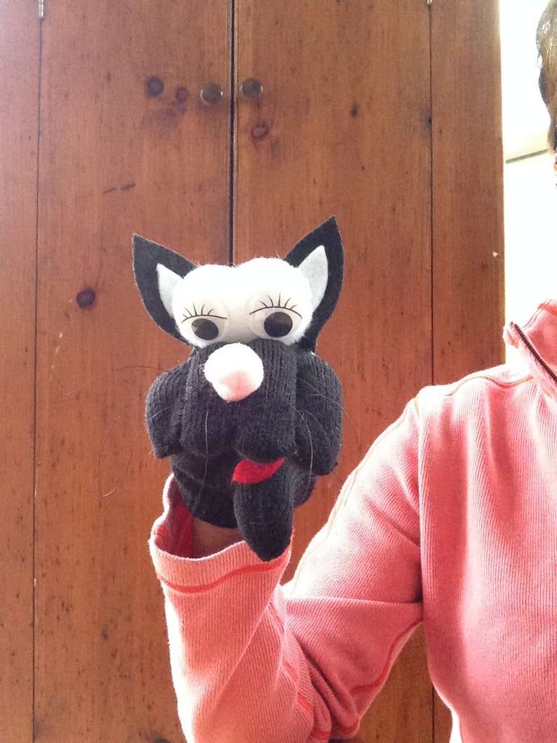 May include: A black and white puppet with a pink nose and large white eyes. The puppet is being held by a person wearing a pink sweater.