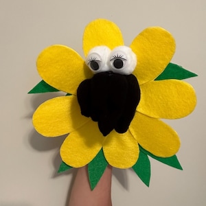 May include: A yellow felt hand puppet shaped like a sunflower with a black felt face, white felt eyes, and green felt leaves.
