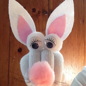 May include: A white sock puppet with pink felt ears and a pink felt nose. The puppet has large googly eyes and a black felt mouth.