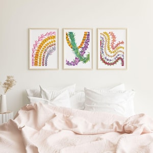 May include: Three framed watercolor prints featuring floral designs in shades of pink, yellow, orange, and purple. The artwork is displayed above a bed with a pink comforter and white pillows. The frames are a light wood color.