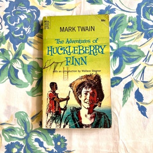 Pode incluir: Um livro de bolso amarelo com uma capa verde que mostra uma ilustração de dois homens em um barco. O título do livro é "As Aventuras de Huckleberry Finn" de Mark Twain. O livro faz parte da série Dell Laurel Edition.