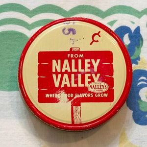 May include: A circular vintage tin with a red and cream design. The tin features the words "NALLEY VALLEY" in bold red letters, with the tagline "WHERE GOOD FLAVORS GROW." The tin has a red rim and is set against a patterned fabric.