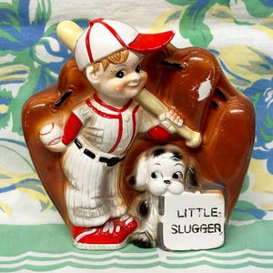 May include: A ceramic figurine featuring a baseball player in a white and red uniform holding a bat, standing in front of a large brown baseball glove. A small white dog sits at the player's feet next to a sign that reads LITTLE SLUGGER.