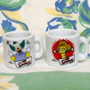 May include: Two small white ceramic mugs featuring characters from The Simpsons. One mug displays Krusty the Clown, the other displays a character with yellow skin and a red dotted background. Both mugs have the show's logo.