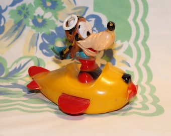 May include: A yellow plastic toy airplane with red wings and a brown and white cartoon dog pilot wearing a blue scarf and a white helmet with a ring on top.