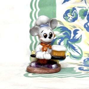 K&ouml;nnte beinhalten: Eine wei&szlig;e Keramikfigur von Mickey Mouse, der als Koch verkleidet ist und ein Tablett mit einem Kuchen darauf h&auml;lt. Der Kuchen ist mit wei&szlig;em Zuckerguss und einer gelben F&uuml;llung verziert. Mickey tr&auml;gt eine wei&szlig;e Kochm&uuml;tze mit einem blauen Streifen, ein wei&szlig;es Hemd mit einem blauen Kragen, eine orangefarbene Fliege und eine violette Hose.