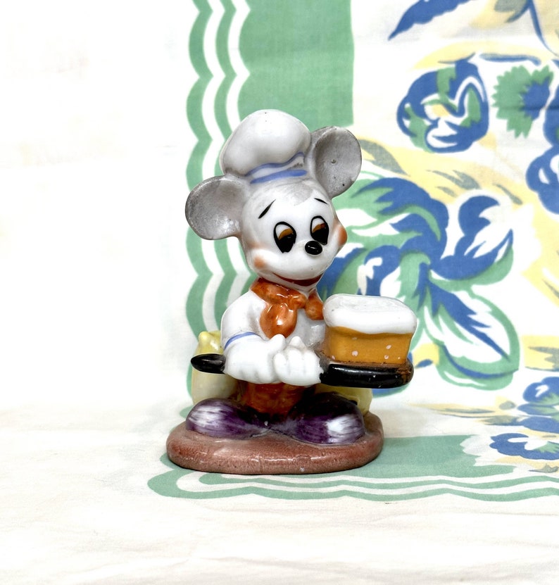 K&ouml;nnte beinhalten: Eine wei&szlig;e Keramikfigur von Mickey Mouse, der als Koch verkleidet ist und ein Tablett mit einem Kuchen darauf h&auml;lt. Der Kuchen ist mit wei&szlig;em Zuckerguss und einer gelben F&uuml;llung verziert. Mickey tr&auml;gt eine wei&szlig;e Kochm&uuml;tze mit einem blauen Streifen, ein wei&szlig;es Hemd mit einem blauen Kragen, eine orangefarbene Fliege und eine violette Hose.