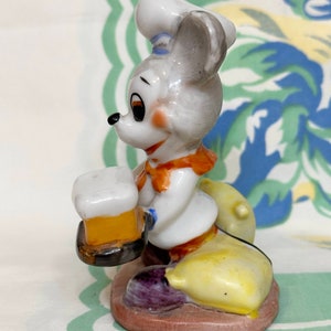 K&ouml;nnte beinhalten: Eine Keramikfigur eines Mause mit einem Kochhut und einem Bierkrug in der Hand. Die Maus sitzt auf einem gelben und violetten Sockel.