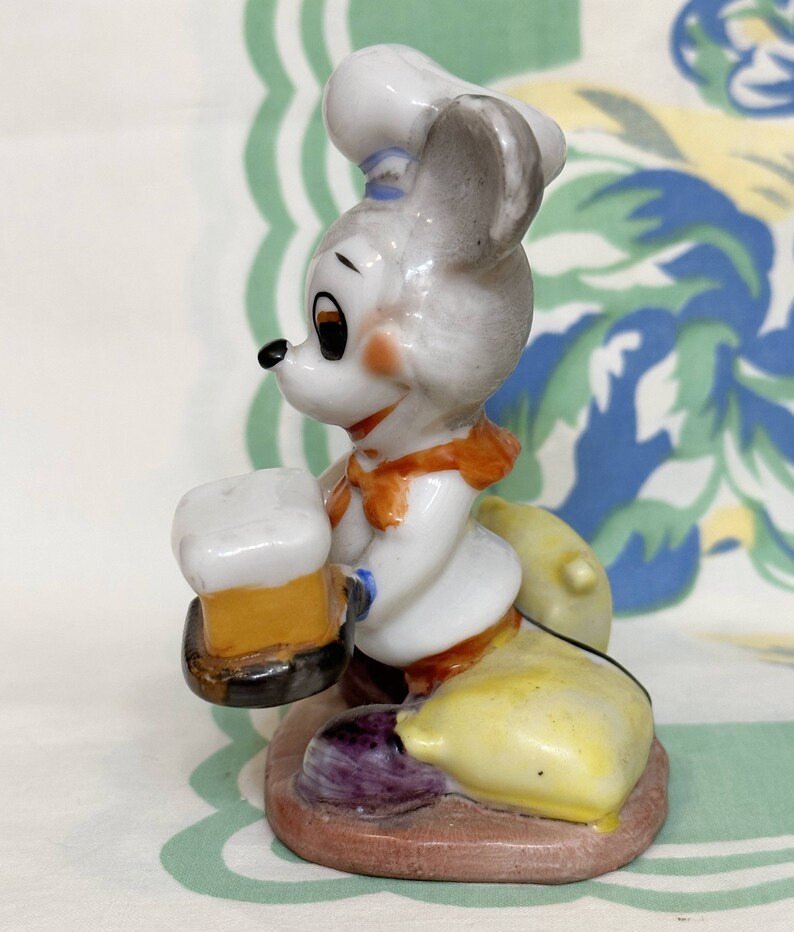 K&ouml;nnte beinhalten: Eine Keramikfigur eines Mause mit einem Kochhut und einem Bierkrug in der Hand. Die Maus sitzt auf einem gelben und violetten Sockel.