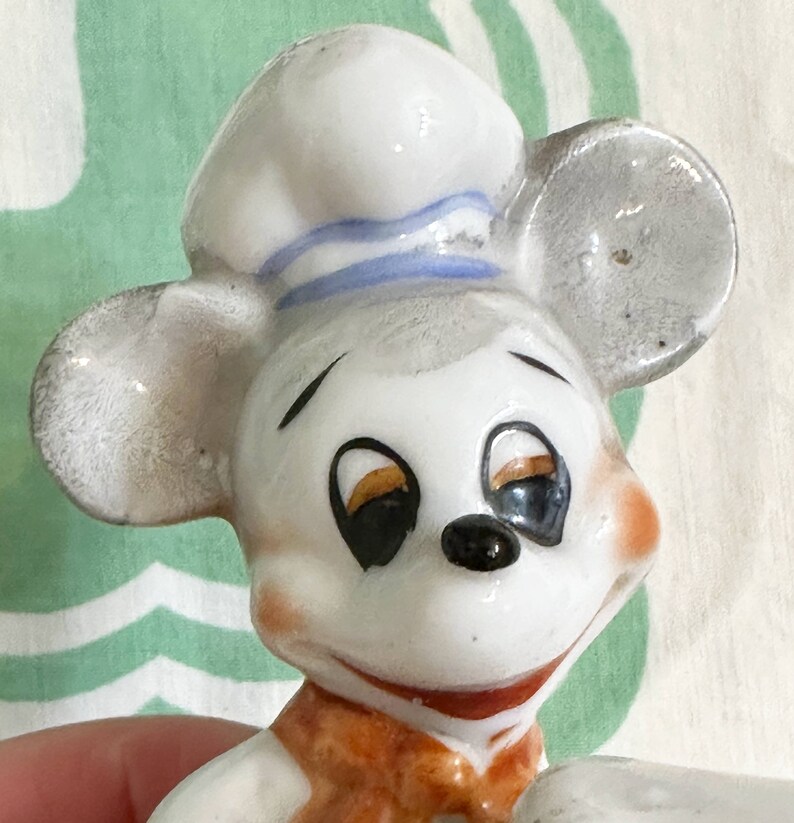 K&ouml;nnte beinhalten: Eine wei&szlig;e Keramikfigur eines Mause mit einer blau-wei&szlig; gestreiften Kochm&uuml;tze. Die Maus hat schwarze Augen, eine schwarze Nase und einen braunen Mund.