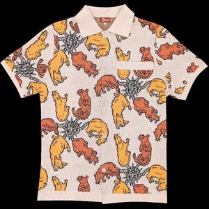 May include: Beige short-sleeved button-up shirt with a repeating pattern of cartoonish brown and yellow animals. The shirt has a cream-colored collar and a small pocket on the chest. The shirt has a unique, eye-catching design.