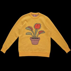 May include: Mustard yellow sweater featuring a graphic of a Venus flytrap plant in a brown pot. The plant has red and green leaves with spiky edges. The sweater has a crew neck and ribbed cuffs and hem.