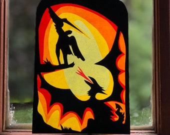 May include: A black paper silhouette of a person riding a dragon against a sunset background. The dragon is flying with its wings spread wide. The sunset is depicted with layers of orange, yellow, and red paper.