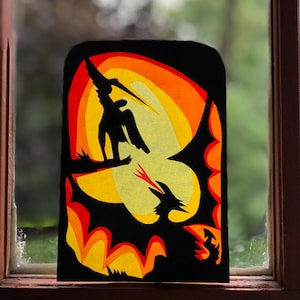 Puede incluir: Una silueta negra de papel de una persona montando un dragón sobre un fondo de puesta de sol. El dragón está volando con sus alas extendidas. La puesta de sol se representa con capas de papel naranja, amarillo y rojo.