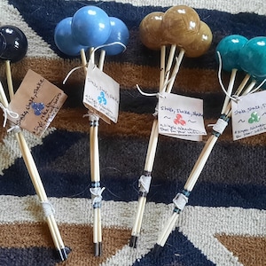 Pode incluir: Quatro conjuntos de chocalhos de madeira com bolas de diferentes cores. Cada conjunto tem três bolas presas a um pau de madeira. As cores são preto, azul, marrom e azul-petróleo.