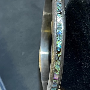 May include: A silver-toned metal bangle bracelet featuring a central inlay of iridescent abalone shell fragments. The bracelet has a smooth, polished finish on the outer edges and a reflective interior surface, showcasing a vibrant mosaic of blue, green, and purple colors.