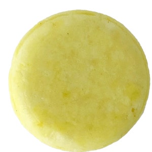 May include: A round, pale yellow solid shampoo bar, viewed from above, against a white background. The bar has a slightly uneven surface and a matte finish. The shape is a flattened disc.