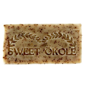 May include: A bar of brown soap with a white label that reads "SWEET ÖKOLE" in a decorative font. The label is surrounded by a wreath of leaves.