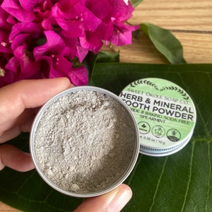 May include: An open, silver-colored tin of herb and mineral tooth powder, with a green and white label. The powder is a light gray color. The label reads "Herb & Mineral Tooth Powder" and "Spearmint". The tin is held near pink flowers and a green leaf.