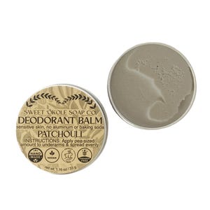 May include: A round metal tin of Sweet 'Ōkole Soap Co. Patchouli deodorant balm with a tan label featuring text, icons, and a net weight of 1.16 oz. The open tin reveals a smooth, light grey, solid balm texture inside the circular container.