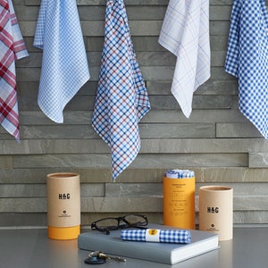 May include: Five different patterned handkerchiefs hanging on a wall, with two brown cardboard tubes containing handkerchiefs and the text "H&G" on the front. The tubes are on a gray surface with a pair of glasses and a blue and white checkered handkerchief.