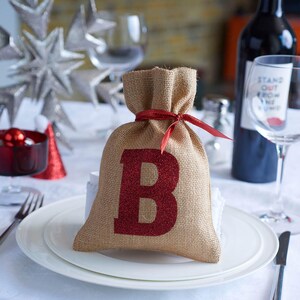 Puede incluir: Una bolsa de regalo de arpillera con una letra "B" roja brillante, atada con una cinta roja. La bolsa está sobre un plato blanco, parte de una mesa festiva con copas de vino y una botella de vino.