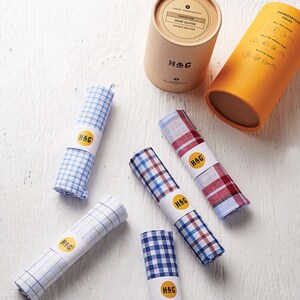 May include: Six different patterned handkerchiefs rolled up and tied with a white band with the text "H&G" in gold lettering. The handkerchiefs are on a white wooden surface.