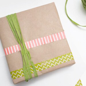 May include: A brown paper gift box wrapped with green and pink patterned washi tape. The box is tied with green shredded paper.
