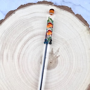 May include: A clear pen with a decorative top featuring two orange fish, green seaweed, and blue accents. The pen has a black ink tip and a clear barrel. The pen is displayed on a wooden slice.