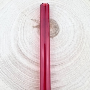 Puede incluir: Una pajita larga y recta de vidrio rojo. La pajita es cilíndrica con un centro hueco. El fondo es una superficie de madera texturizada de color marrón claro con anillos de crecimiento circulares.