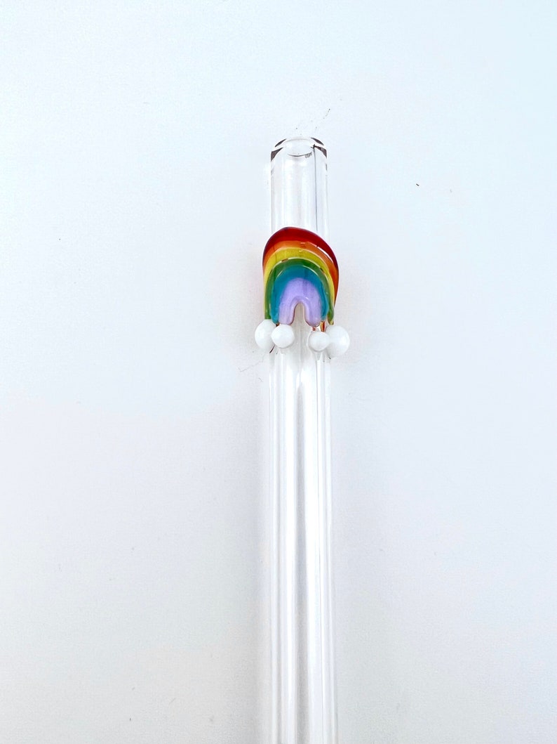 May include: A clear glass drinking straw featuring a decorative fused glass rainbow accent. The rainbow is composed of red, orange, yellow, green, blue, and purple bands, with small opaque white glass spheres attached at the base of the rainbow.
