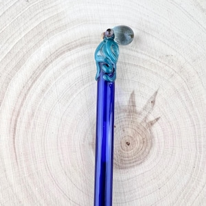 May include: A handcrafted glass pen with a blue body and a teal octopus design at the top. The pen features a clear, rounded tip and is set against a light wood grain background. The octopus design adds a unique, artistic touch.