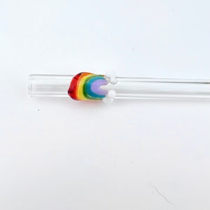 May include: A clear glass drinking straw featuring a decorative glass attachment shaped like a rainbow with small white clouds on either side. The rainbow is composed of red, orange, yellow, green, blue, and purple bands of colored glass.