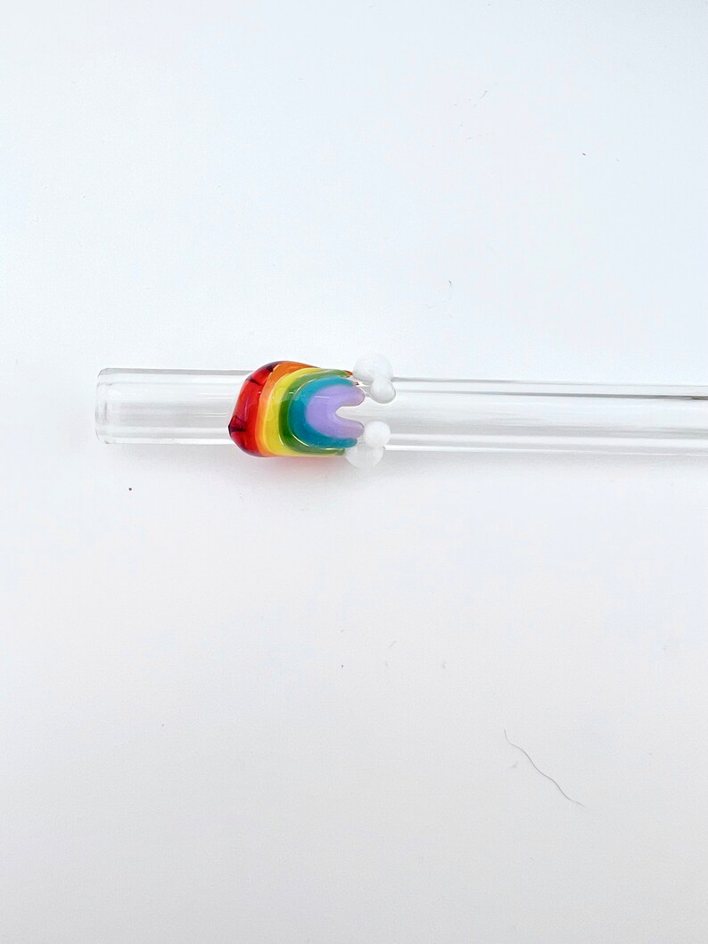 May include: A clear glass drinking straw featuring a decorative glass attachment shaped like a rainbow with small white clouds on either side. The rainbow is composed of red, orange, yellow, green, blue, and purple bands of colored glass.