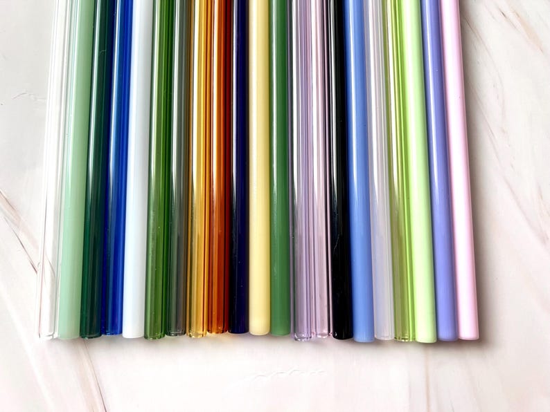 May include: A collection of colorful glass straws in various shades, including clear, green, blue, yellow, and purple. The straws are arranged vertically, showcasing their smooth, cylindrical shape. These reusable straws offer an eco-friendly alternative to plastic.