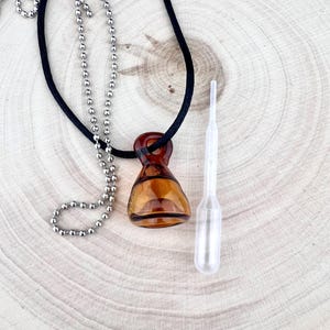 May include: A brown glass pendant necklace with a black cord and a silver ball chain. A clear plastic dropper is next to the pendant. The pendant has a unique shape and is made of amber-colored glass.