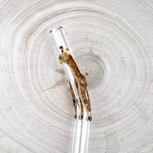 Puede incluir: Una pajita para beber de vidrio transparente con un diseño de jirafa. La jirafa es marrón y beige con manchas negras.