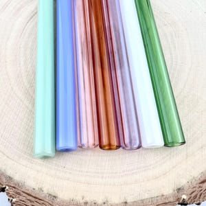 以下が含まれることがあります： 緑、青、ピンク、茶色の色合いのカラフルなガラスストローのコレクション。 ストローは、明るい茶色の質感のある木製の表面に配置され、鮮やかな色合いと滑らかな円筒形のデザインを示しています。
