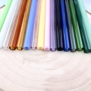 May include: A collection of colorful glass straws in various shades, including green, white, amber, blue, yellow, and purple. The straws are arranged on a light-colored wooden surface, showcasing their vibrant hues and transparent quality.