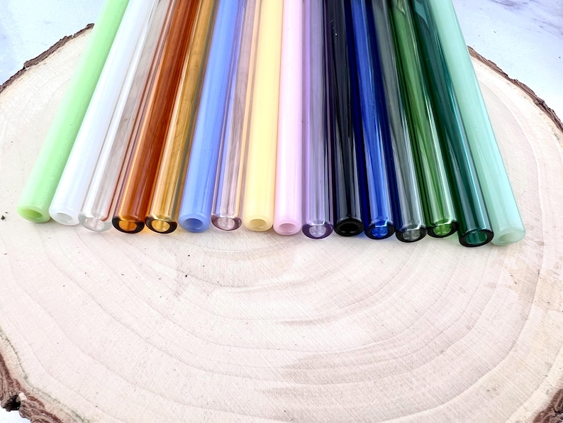 May include: A collection of colorful glass straws in various shades, including green, white, amber, blue, yellow, and purple. The straws are arranged on a light-colored wooden surface, showcasing their vibrant hues and transparent quality.