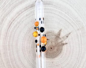 Può includere: Una cannuccia in vetro trasparente decorata con una serie di dettagli sferici in rilievo in vetro nero solido, arancione opaco e giallo traslucido, disposti a grappolo lungo la sezione centrale del corpo cilindrico della cannuccia.