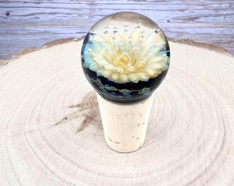 May include: A bottle stopper featuring a spherical glass topper with a clear exterior and a central yellow flower-like design encased in dark blue glass. The glass sphere is mounted on a tapered, light-colored natural cork base.