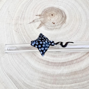 May include: A glass stingray figurine featuring a black body with white polka dots and a long, slender, curved tail, attached to a clear, cylindrical glass rod. The stingray has a flat, diamond-shaped body with a smooth, glossy finish.