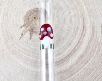 Può includere: Una cannuccia in vetro trasparente con un fungo decorativo rosso a pois bianchi e gambo bianco, arricchita da piccole linee verticali verdi alla base del disegno, creando un accessorio in vetro dettagliato e colorato per le bevande.