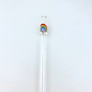 May include: A clear glass drinking straw featuring a small, colorful rainbow decoration near the top. The rainbow is composed of red, orange, yellow, green, blue, and purple stripes, with two small white glass spheres attached at each base.