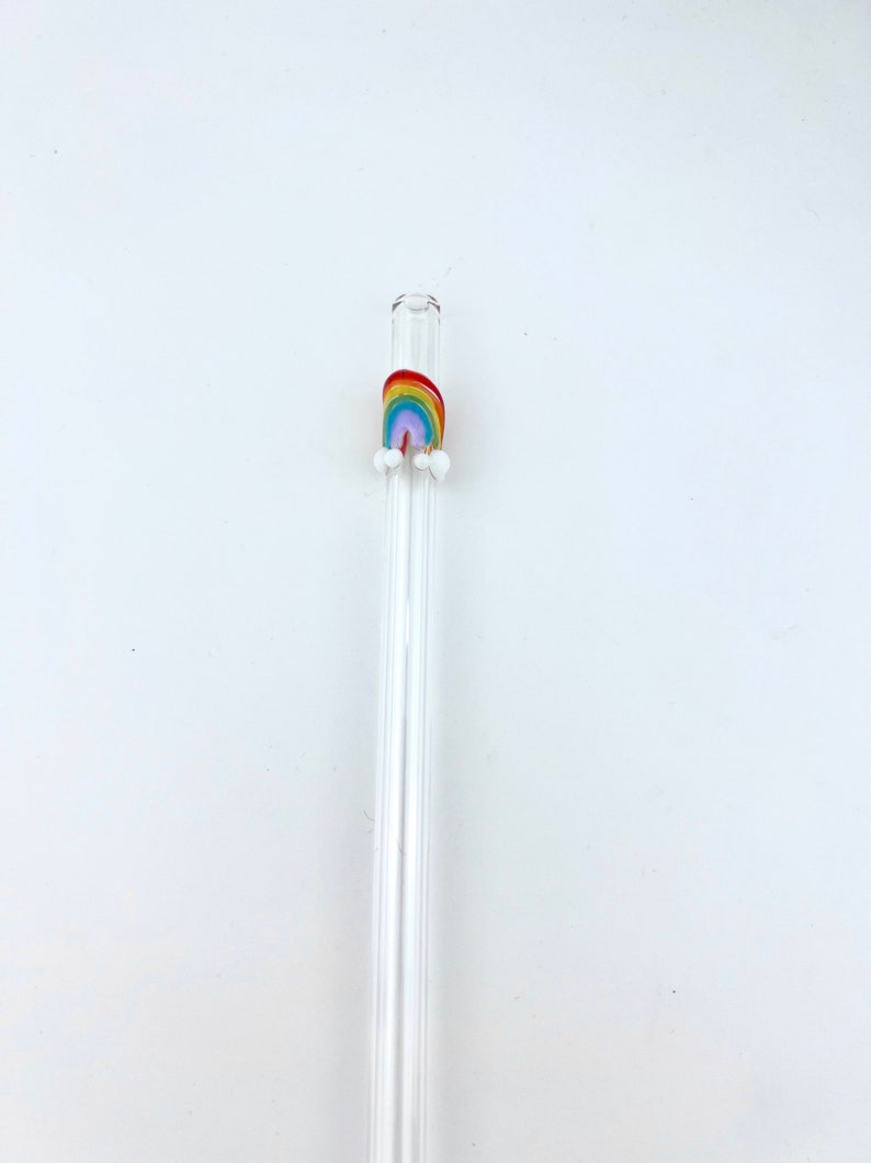 May include: A clear glass drinking straw featuring a small, colorful rainbow decoration near the top. The rainbow is composed of red, orange, yellow, green, blue, and purple stripes, with two small white glass spheres attached at each base.