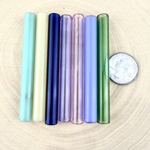 May include: A collection of seven glass straws in various colors, including light green, yellow, navy blue, pink, purple, light blue, and dark green. A U.S. quarter is next to the straws for size comparison. The straws are cylindrical and appear to be handmade.