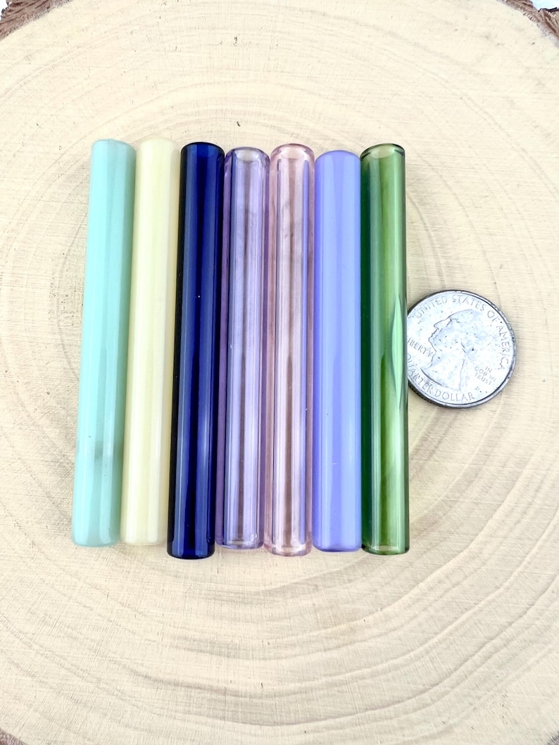 May include: A collection of seven glass straws in various colors, including light green, yellow, navy blue, pink, purple, light blue, and dark green. A U.S. quarter is next to the straws for size comparison. The straws are cylindrical and appear to be handmade.